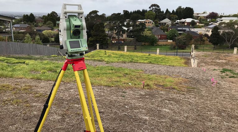 Surveying instrument on a tripod set up on a vacant residential block, with survey pegs marking the site